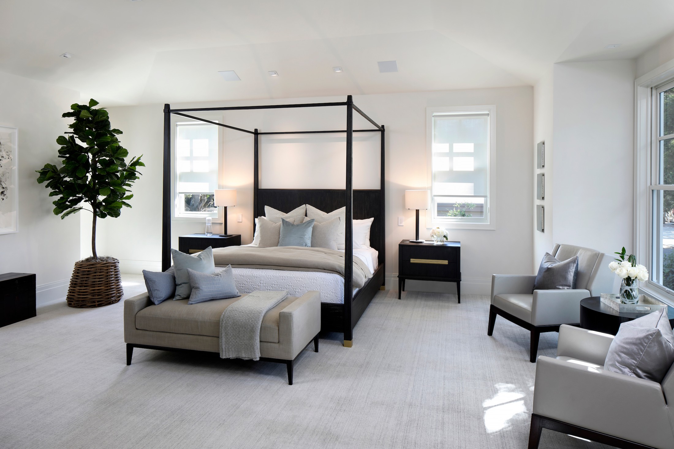 Master bedroom with black canopy bed, ottoman, seating area, and a large tree in the corner of the space.