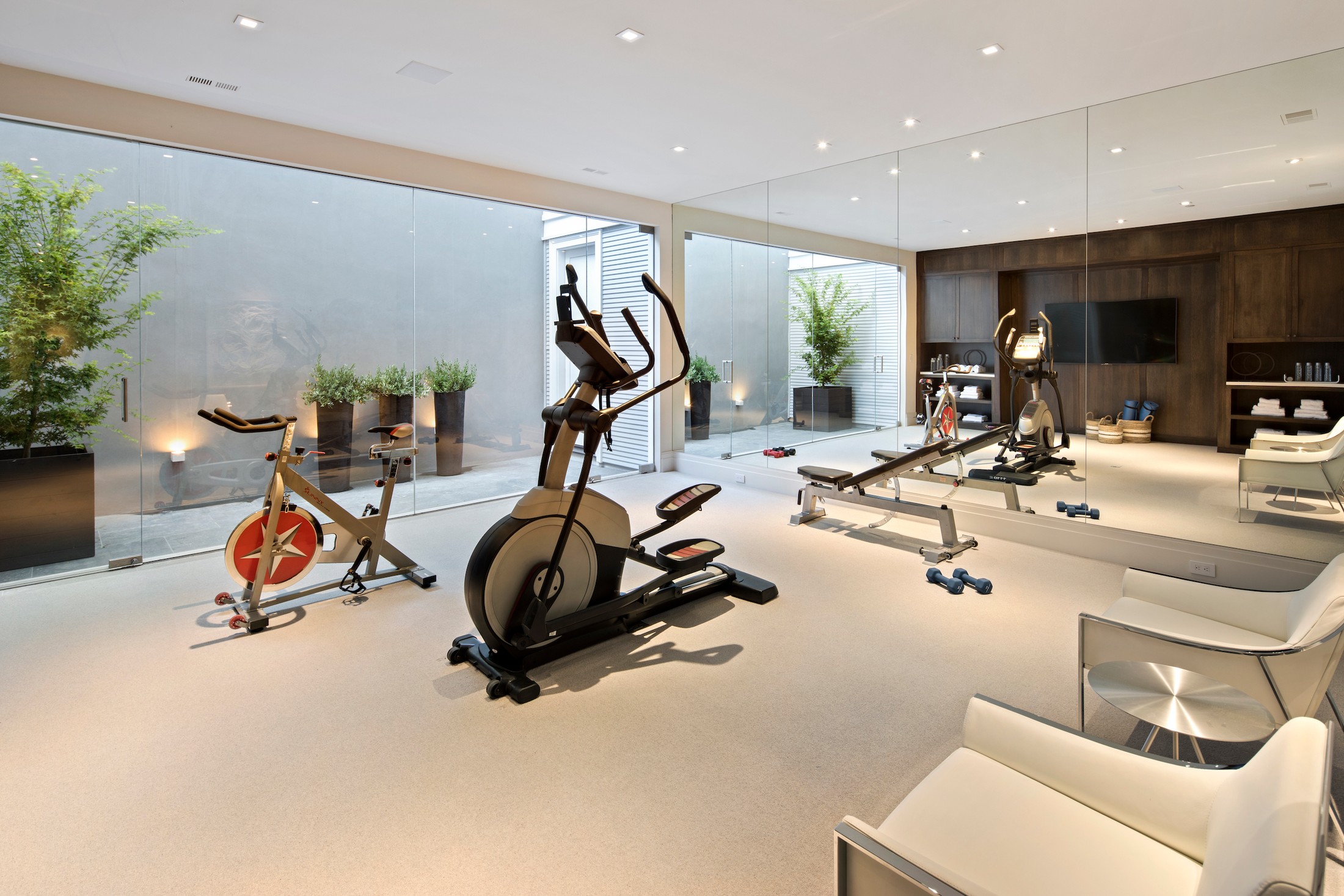 Gym area with different bike machines, a seating area, and there are large windows which lead out to different trees planted in pots.
