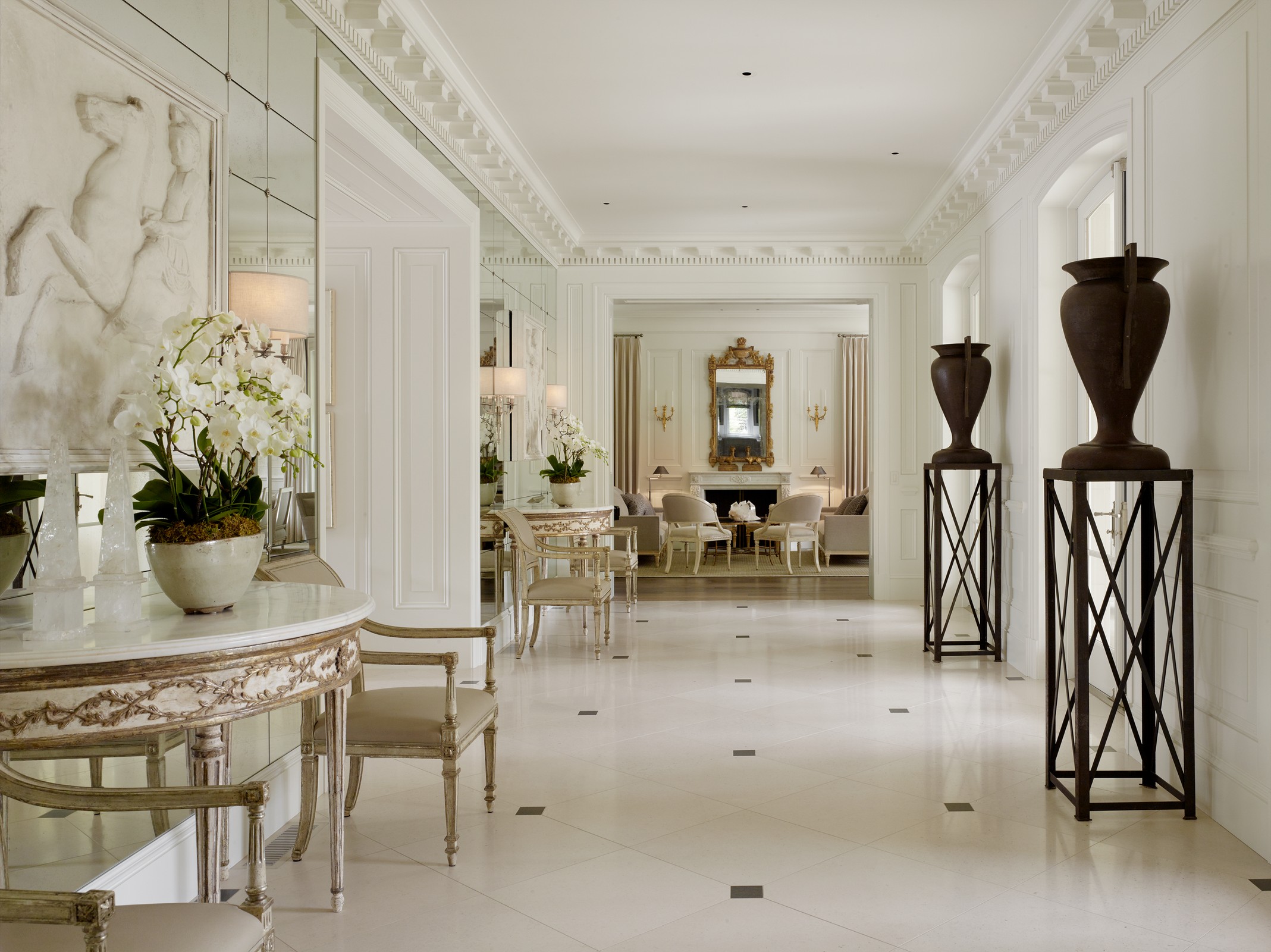 Hallway which leads into a living room. There are large black pots on stands and a white table with flowers on it with flower detailing on the table