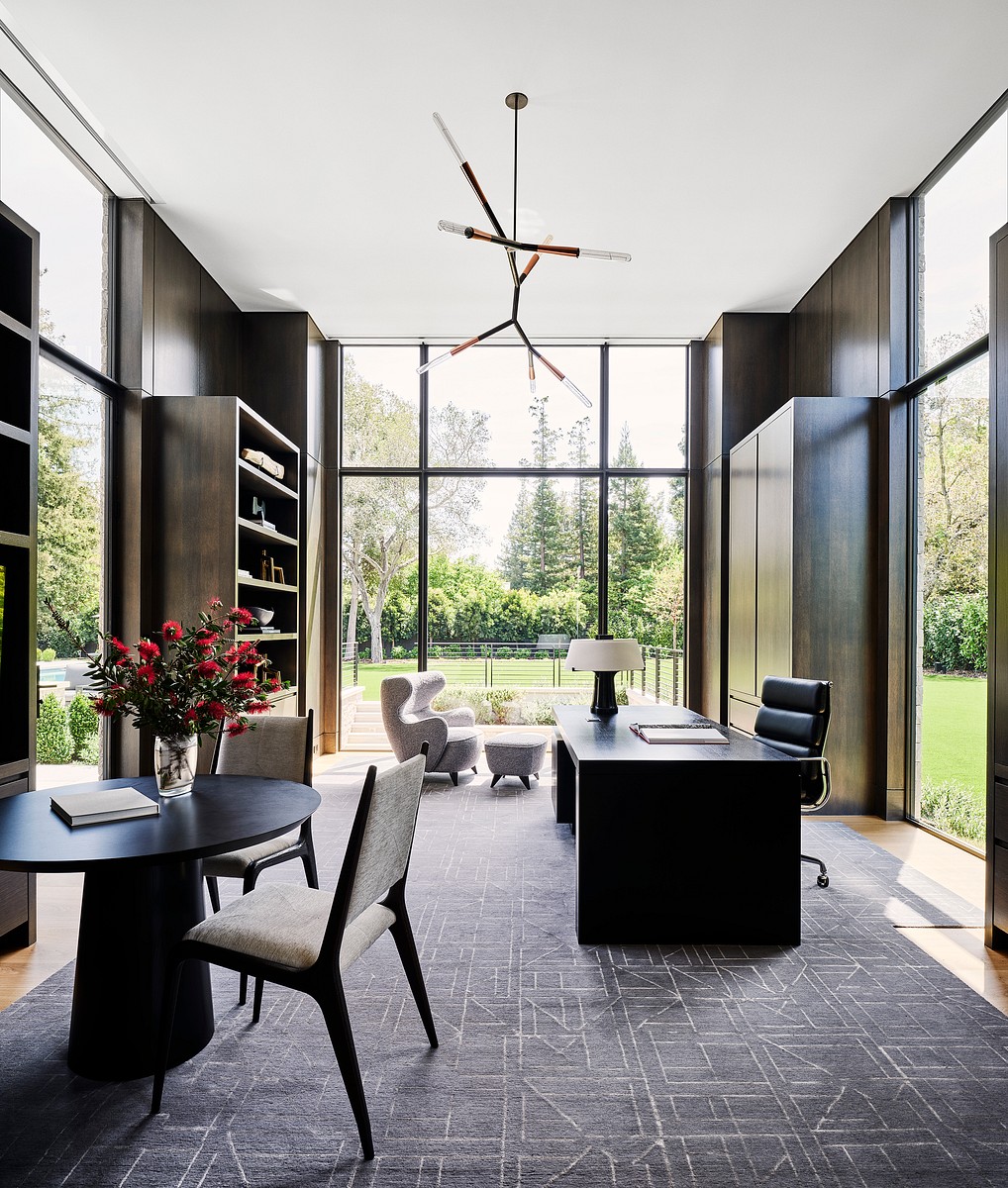 Office space with patterened rug, and a black desk with multiple seating area and wooden shelves.