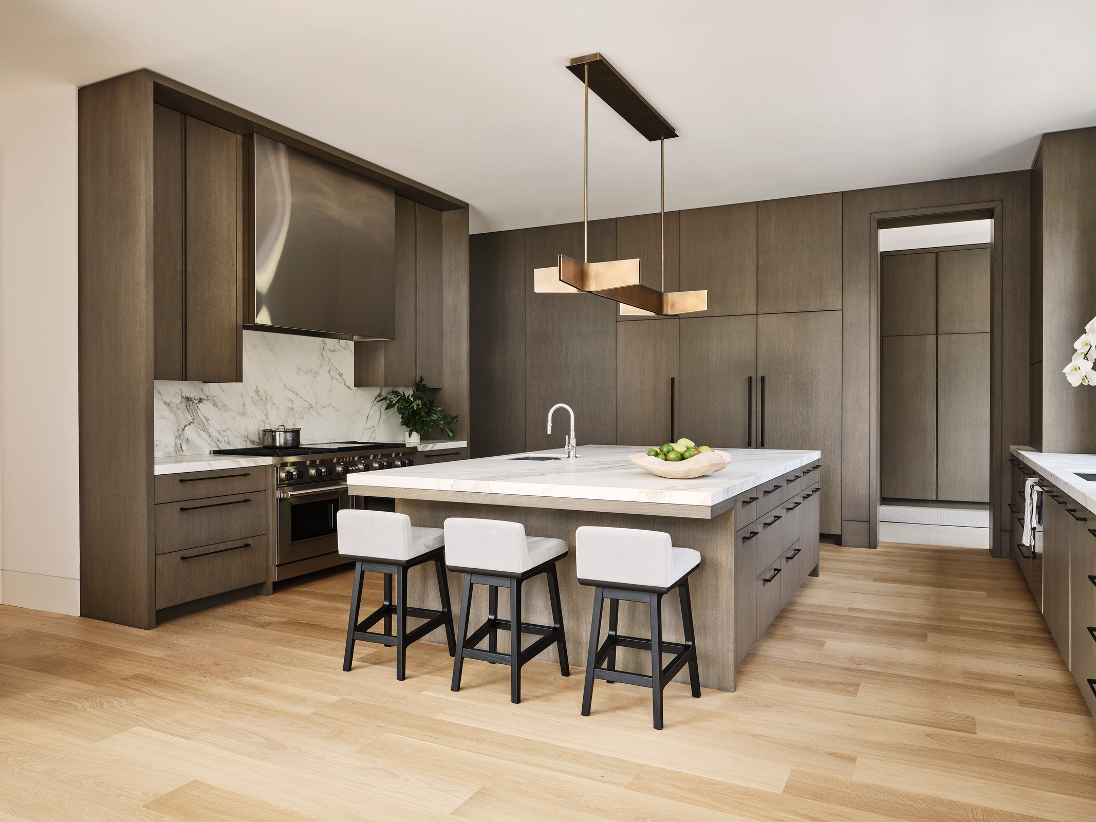 Kitchen which has dark wood and brass detailing on the lamps. There is a large island in the center and white/black chairs.
