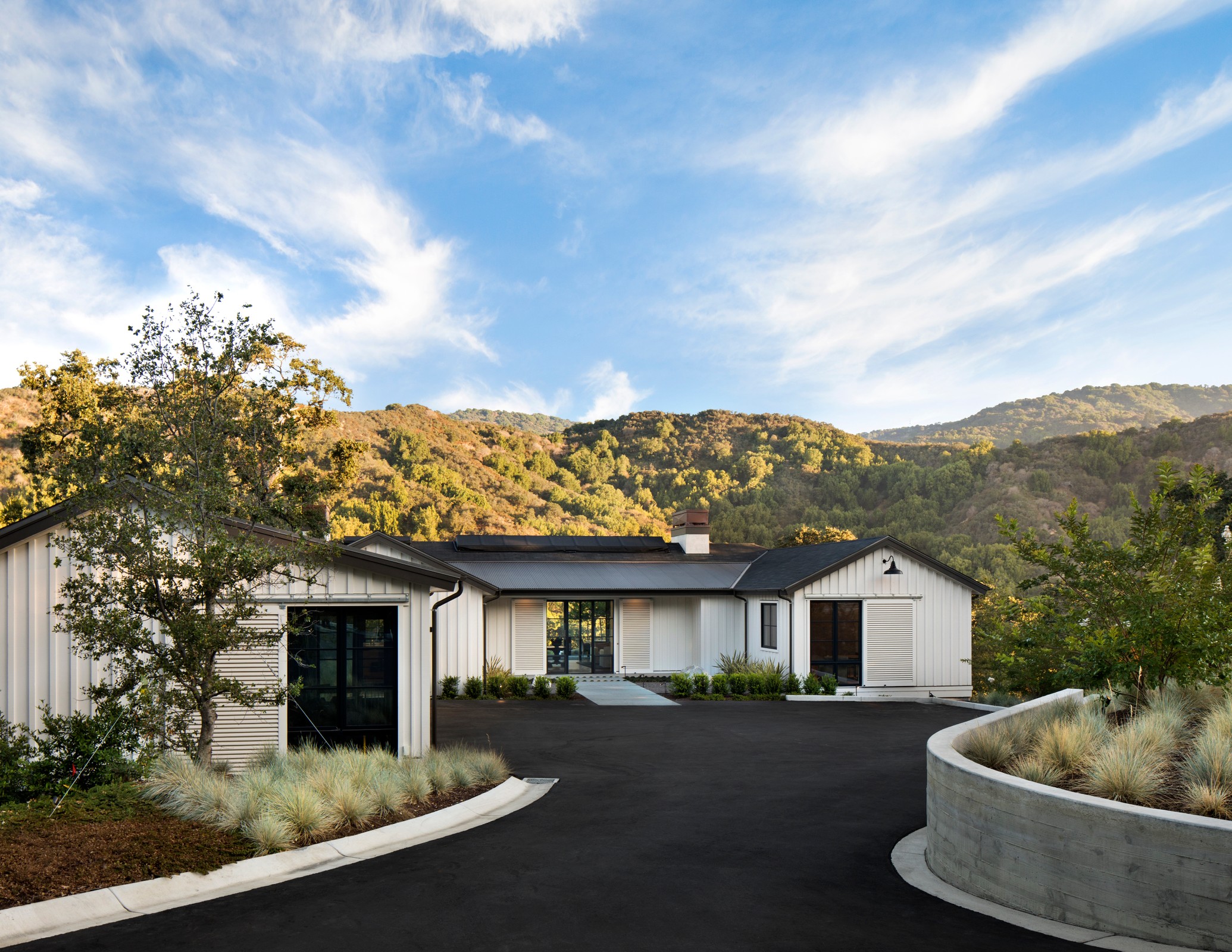 Exterior image of the front of the home, black driveway which is curvy. The home is white with vertical siding. Hills with trees in the background.