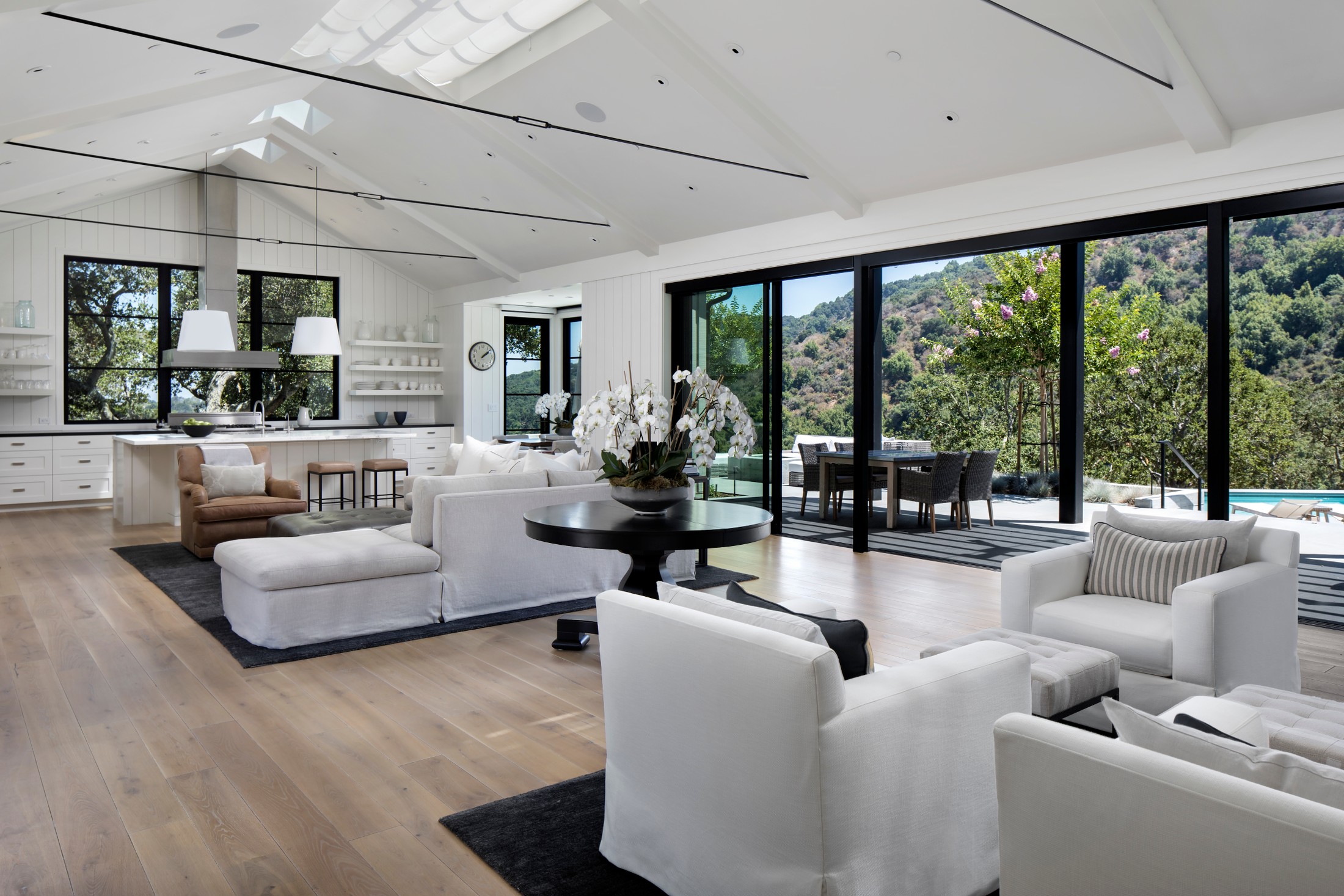 Living room area with vaulted white ceilings, neutral colored furniture, black table with white flowers on it, big rectangular doors which go to an outdoor backyard.