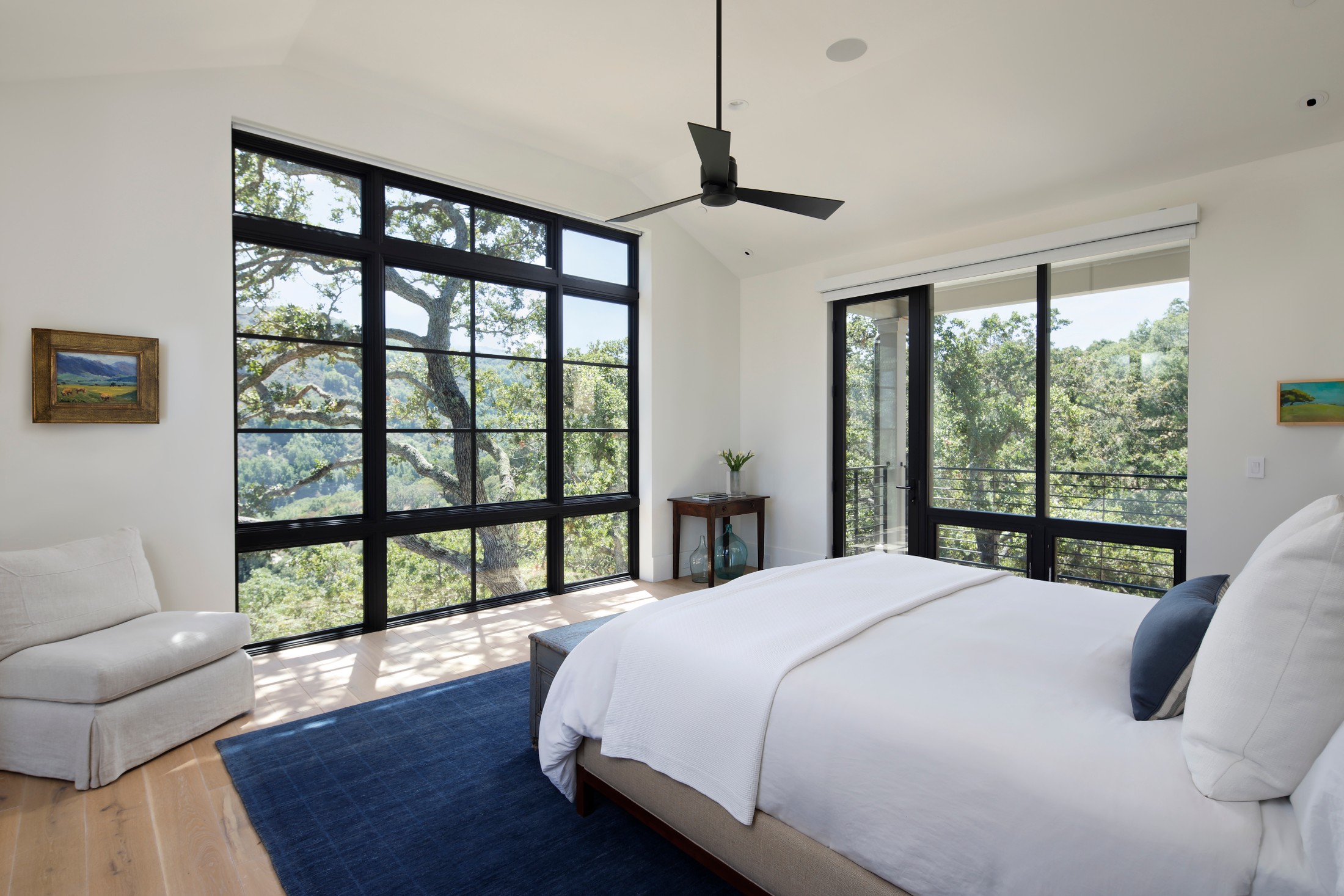 Bedroom with a black ceiling fan, windows with black detailing, wooden floors, and oatmeal colored bedding and furniture. Small piece of artwork on the wall of a landscape with wooden frame.