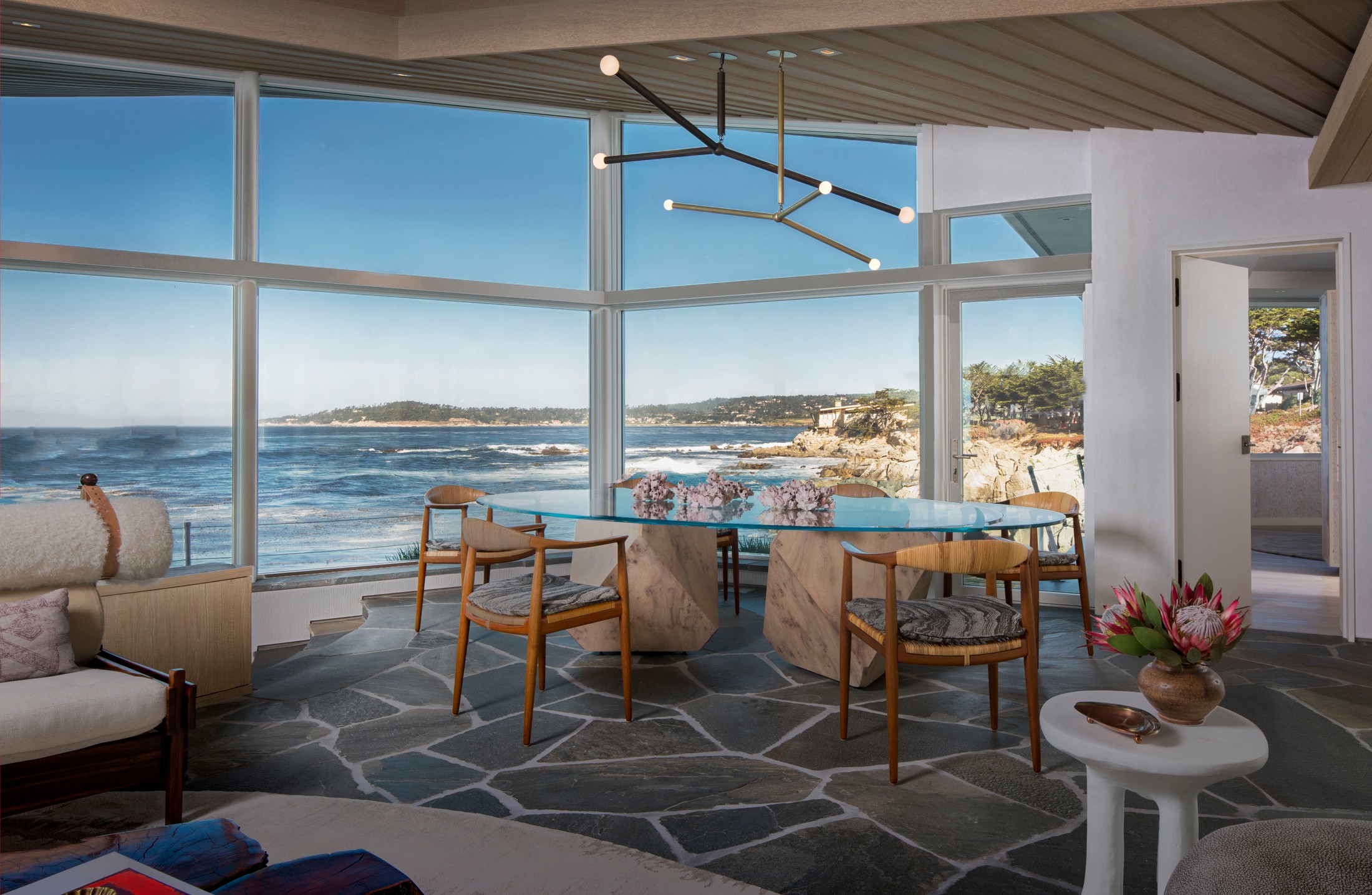 Dining room, with glass table with stones supporting the table below. Wooden chairs and stone flooring. Very large windows floor to ceiling with the ocean in the background.