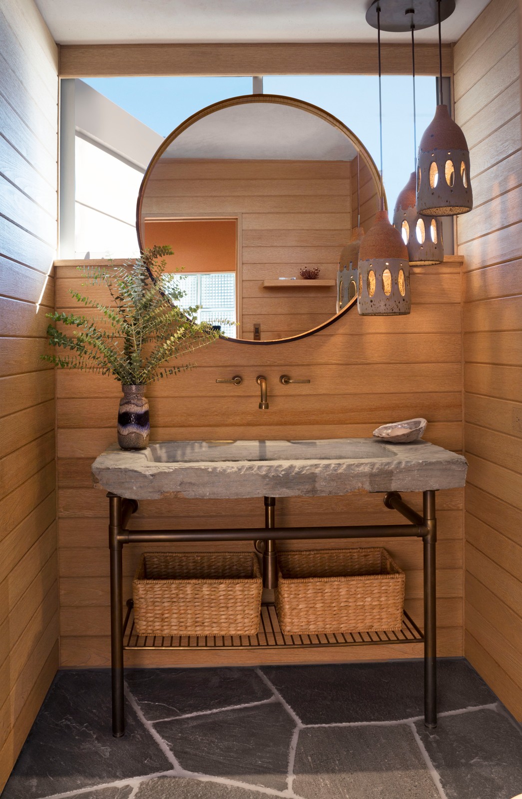 Powder room with a circular mirror, clay hanging lamps from the ceiling, stone sink, and flowers in a vase.