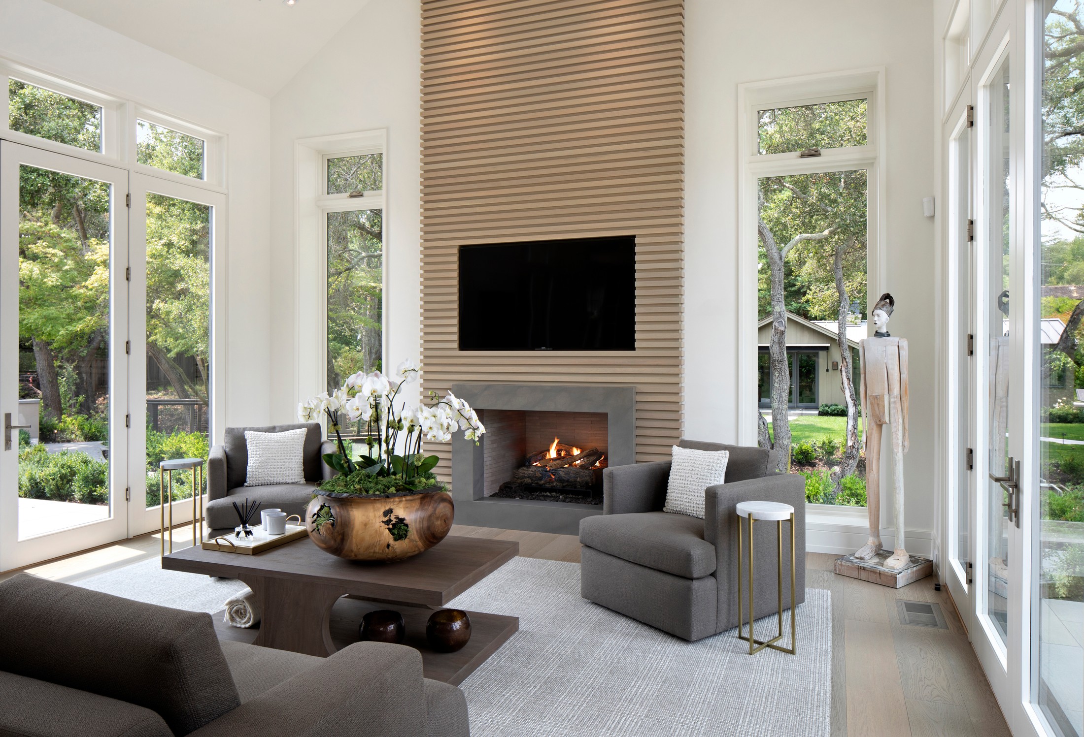 Family room with a large tv on the wall above a lit fireplace. There is a sculpture in the corner of the room. There are large floor to top of the wall windows surrounding the space. Couch, chairs and a table sit in the center.