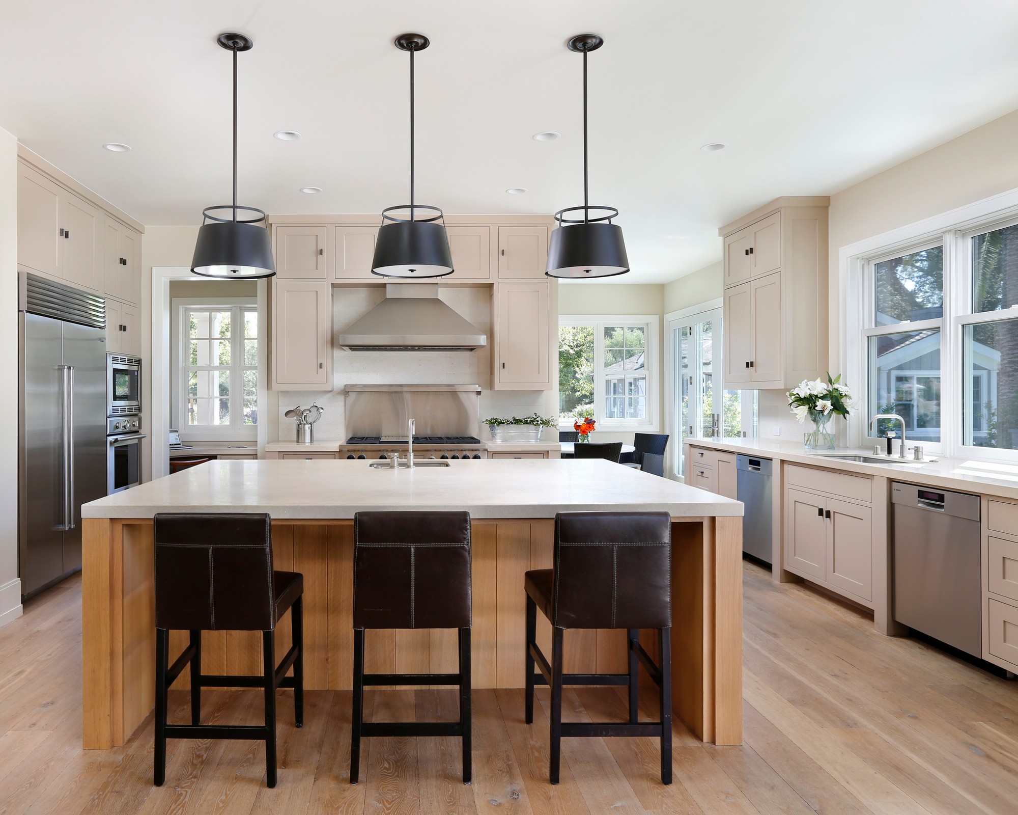 Kitchen with large black lamps hanging from the white ceiling. There are threee black chairs and a large island in the center with a sink.