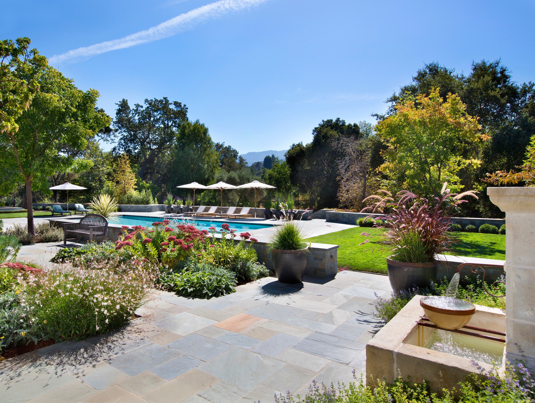 Outdoor pool, with lounge chairs and umbrellas. Landscaped sections with flowers and plants. The pool is surrounded by trees and hills are seen from a far. There is a small fountain on the right side