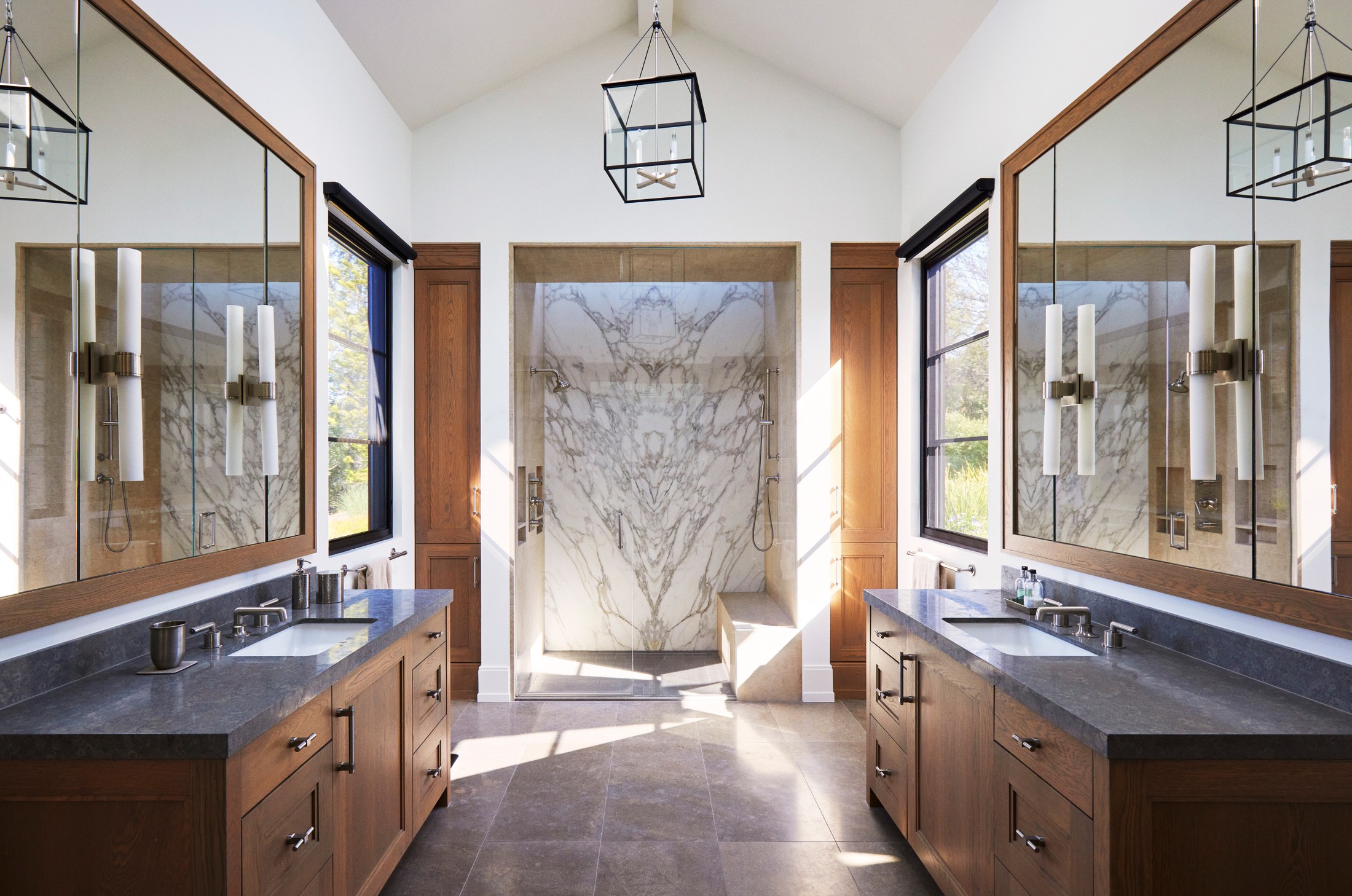 Image of a bathroom. On each side you can see sinks and vanity areas with mirrors. In the center of the image in the distance, is a marble shower. There is a black lamp hanging from the ceiling.