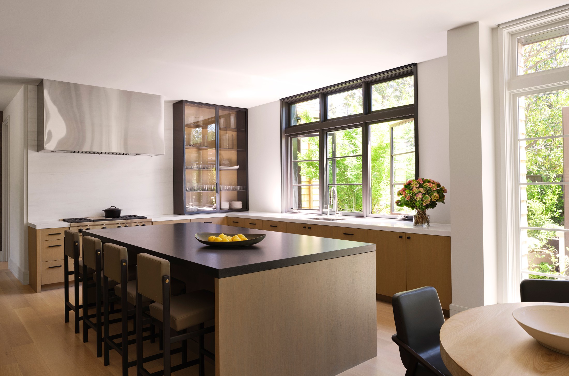 Image of a kitchen, which includes yellow flowers on the countertop. There is lemons in a tray on the countertop, as well. Wooden cabinets and black detailing on the windows.