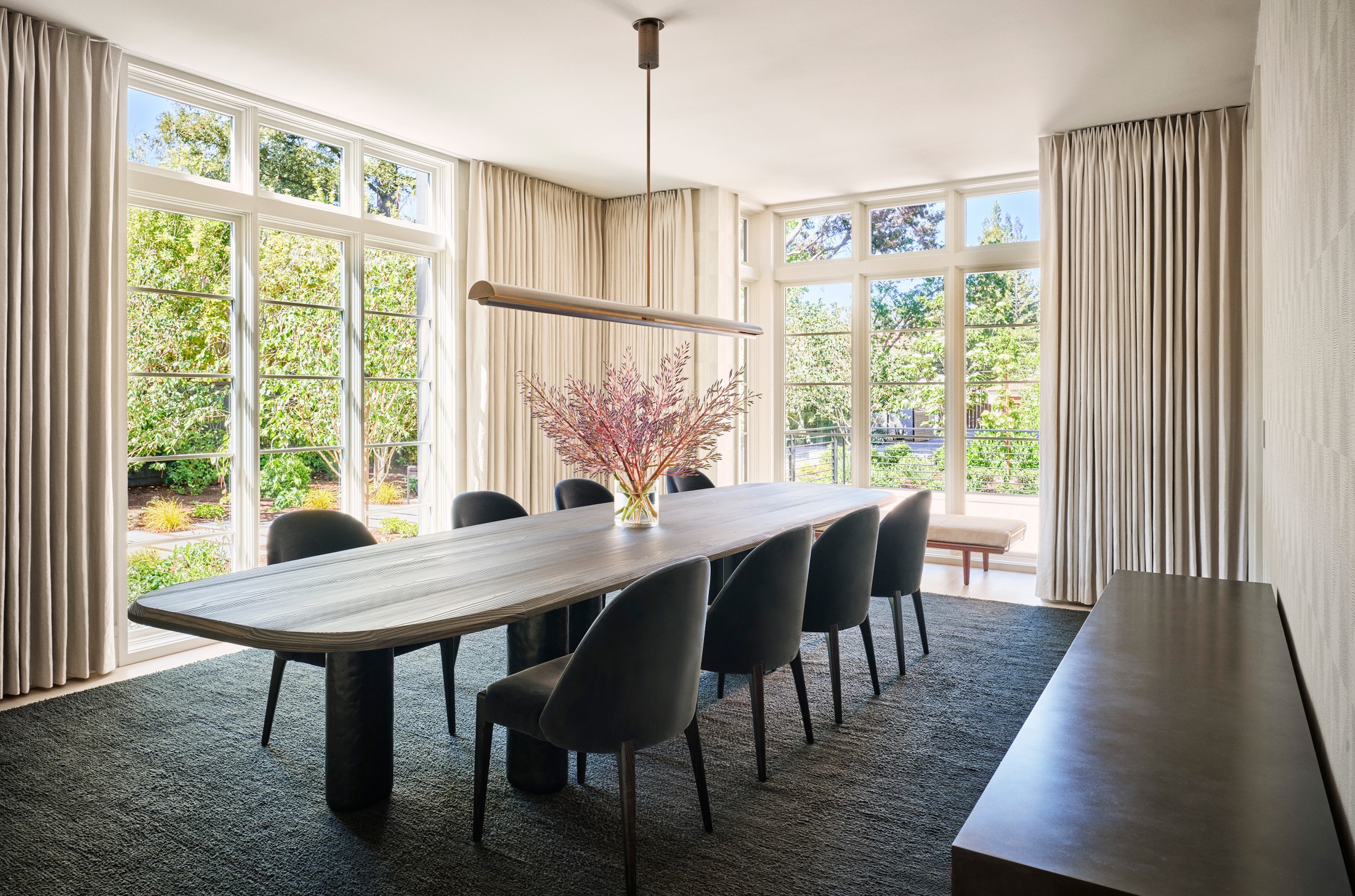 Image of a dining room, with large white windows. There are circular chairs and a large rectangular table with flowers in the center of the table.