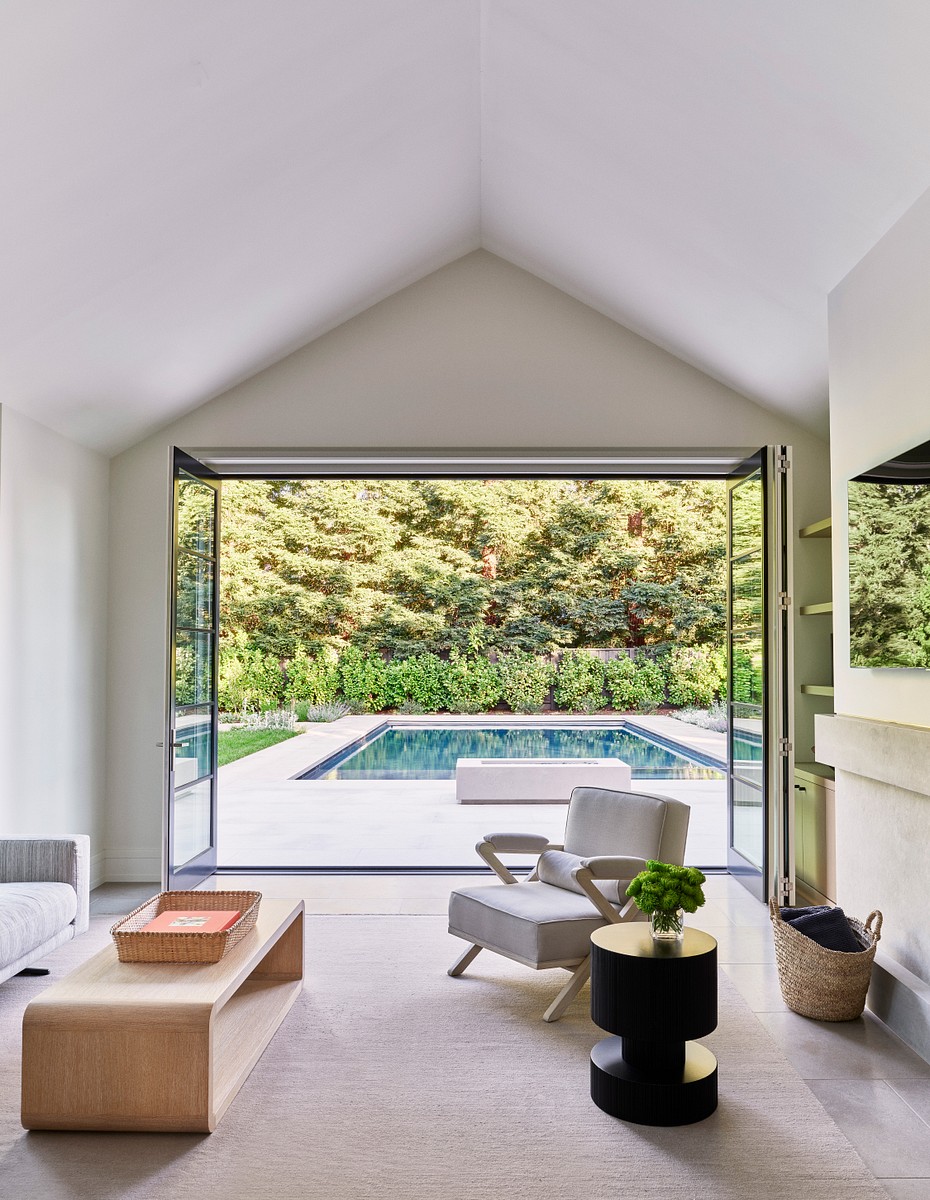 Image of a room with triangular ceilings, chairs, wooden table, black nightstand, flowers, a woven towel bag, and very large doors which lead to the outside with a pool.