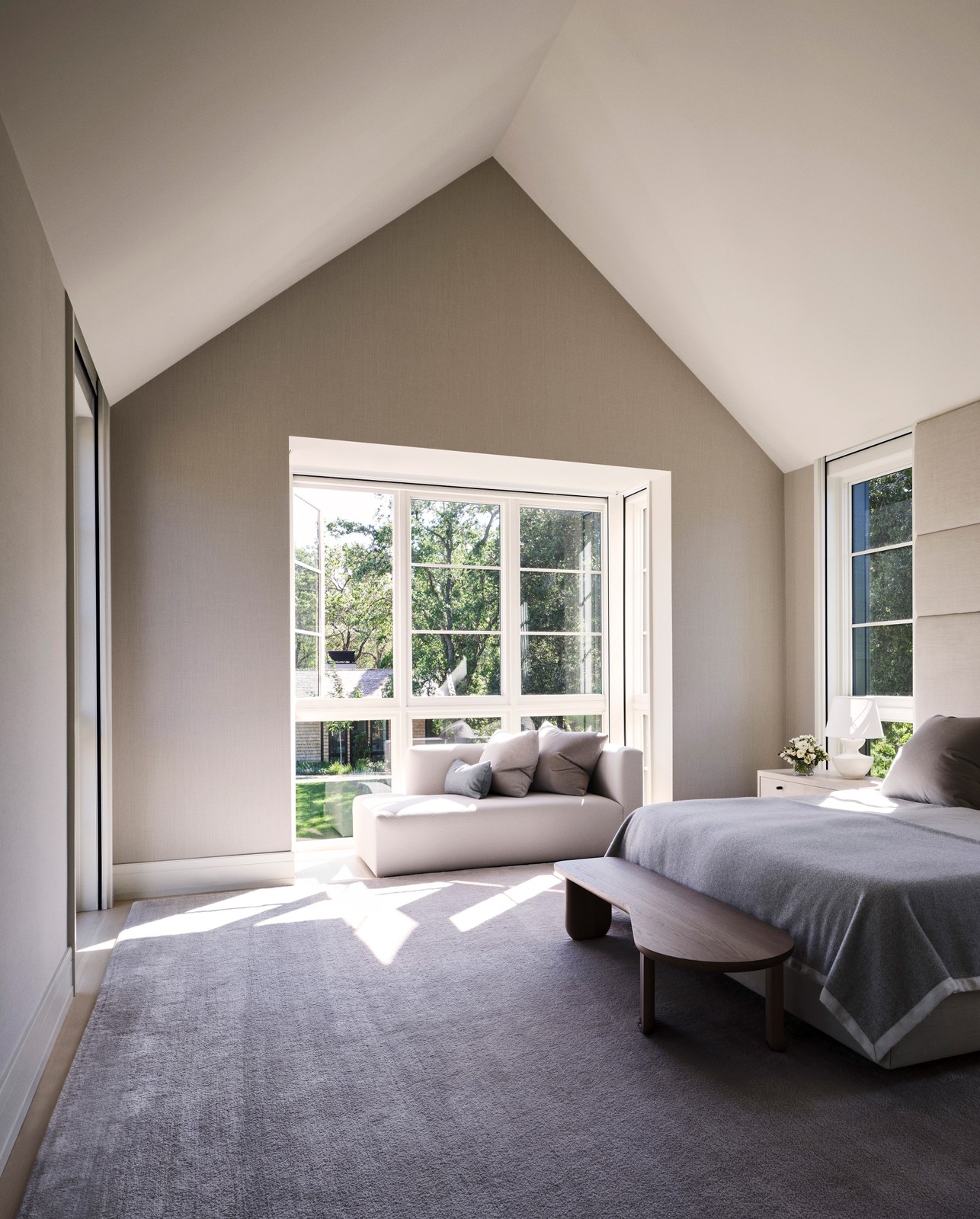 Image of a bedroom, with triangular ceilings and white windows. There are flowers on the nightstand and a white lamp.